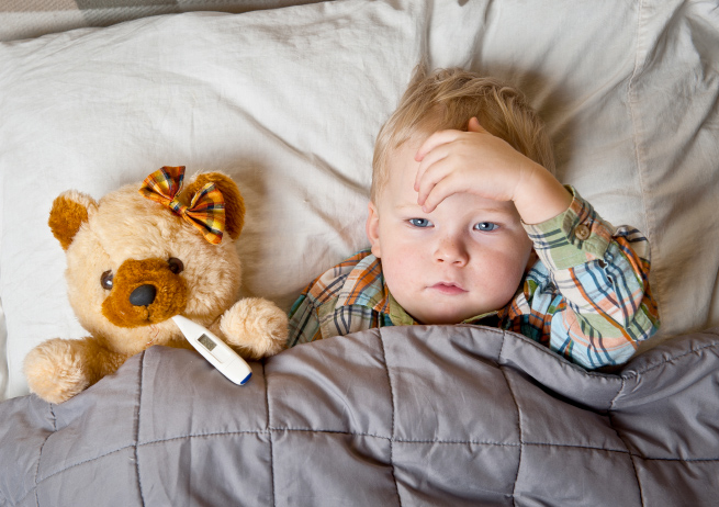 Child laying next to a teddy bear with a thermometer in bed